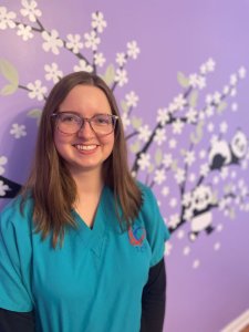 Emily, neurodiversity-affirming speech therapist, is a caucasian female with glasses and shoulder-length straight light brown hair. She is smiling and standing in front of a purple wall with panda and tree decal decorations in a therapy room.