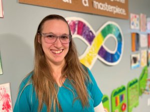 Emma Neurodiversity-Affirming Occupational Therapist, is a caucasian female with long straight blond hair standing, smiling while standing in front of a rainbow infinity symbol on the wall behind her.