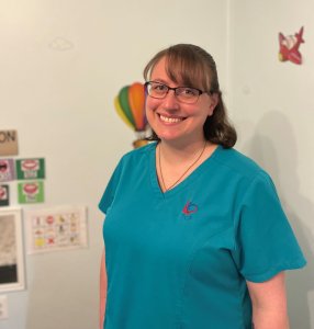 Megan, a neurodiversity-affirming speech therapist, is a caucasian female with bangs and glasses, with shoulder-length light brown hair. She is smiling and standing in front of a light blue wall with therapy posters in the background, in a treatment room.