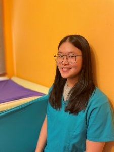 Marissa, neurodiversity-affirming occupational therapist, is an Asian female smiling in front of an orange wall and next to a piece of therapy equipment made out of lycra.