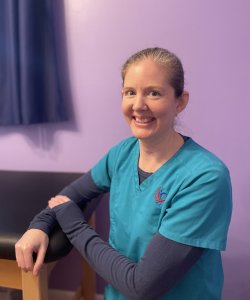 Andrea, neurodiversity-affirming occupational therapist, is a caucasian female with hair pulled back, smiling while one arm rests on black treatment table.