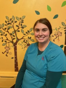 Brenna, neurodiversity-affirming occupational therapist, is a caucasian female with brown hair pulled back, smiling while sitting in front of an orange wall with monkey decal decorations in a pediatric therapy room