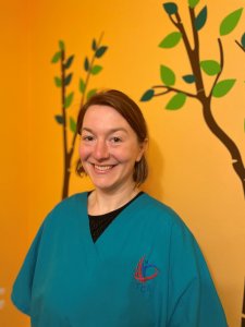 Sophie, neurodiversity-affirming speech therapist, is a caucasian female with short light brown hair. She is smiling and standing in front of an orange wall with tree decals in a therapy room